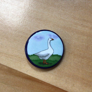 A round button with an illustrated goose walking in the grass. 