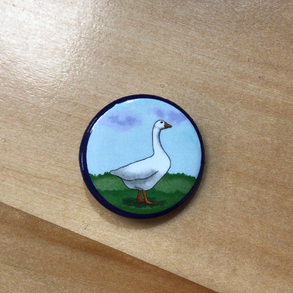 A round button with an illustrated goose walking in the grass. 