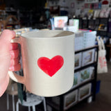 A ceramic mug with a bright reddish pink heart printed in the middle is held up from the left side of the image. 