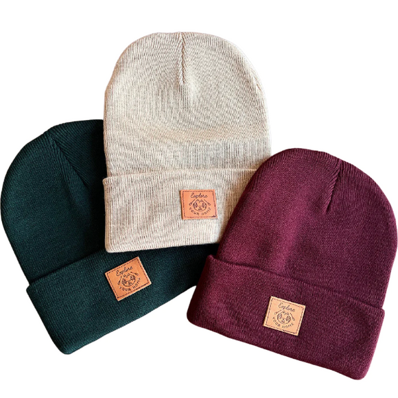 Three toques lay flat on a white background, green, beige and burgundy. All have a square leather patch reading 