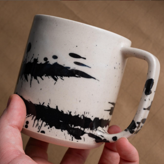 Off-white ceramic mug with handle and black paint splotch design. 