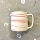 Unglazed mug with uneven pink stripes wrapping around it over an off-white background. 
